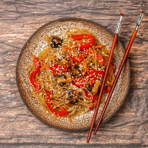 Starch (rice, potato) noodles with mushrooms and vegetables-bell peppers, carrots, onions, sesame seeds and soy sauce. Vegetarian dish. Delicious dinner in Asian (Chinese, Korean, Japanese) style. Selective focus, square picture fideos de almidón (arroz, patata) con champiñones y verduras-pimientos, zanahorias, cebollas, semillas de sésamo y salsa de soja. plato vegetariano. deliciosa cena en estilo asiático (chino, coreano, japonés). enfoque selectivo, imagen cuadrada