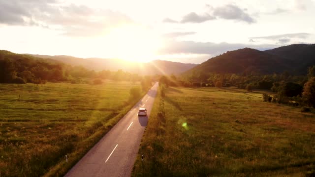 A gray car drives down an empty country road at a golden summer sunset