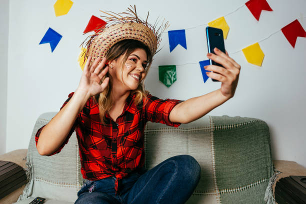 Brazilian Festa Junina at home. Person wearing typical clothes. Living room decorated with colorful flags. Woman using cellphone. Brazilian Festa Junina at home. Person wearing typical clothes. Living room decorated with colorful flags. Woman using cellphone. straw hat photos stock pictures, royalty-free photos & images