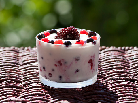 Close-up of a glass bowl of white yogurt with fresh berries on a rattan garden table in sunlight