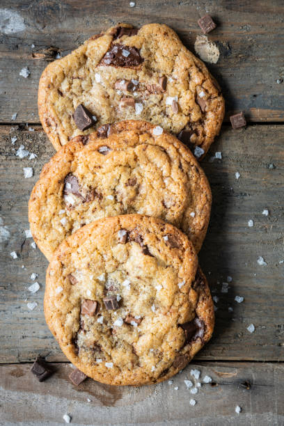 Three freshly baked chocolate chip cookies on a wooden background Three freshly baked chocolate chip cookies are lying on a textured wooden background sprinkled with salt. There are loose chocolate chips and salt flakes scattered around the cookies. The chocolate in the cookies has melted leaving the biscuits gooey. Taken from overhead. chocolate chip cookie stock pictures, royalty-free photos & images