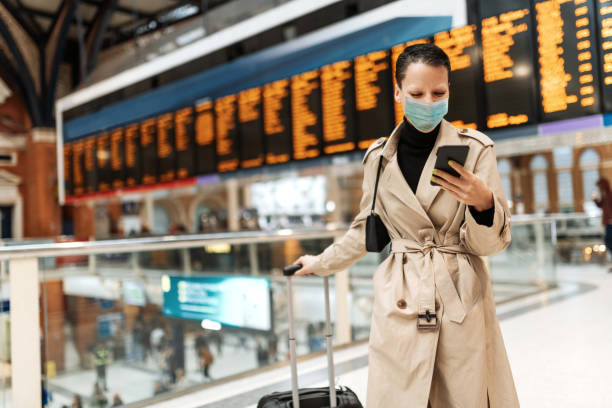 podstawowe podróże podczas blokady - kobieta z maską na twarz melduje się online, czekając w pobliżu tablicy odlotów - arrival departure board airport travel business travel zdjęcia i obrazy z banku zdjęć