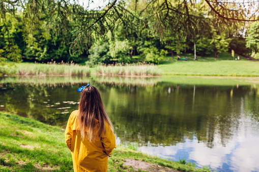 Young woman in yellow raincoat walking in summer park. Girl admires lake landscape view. Enjoying nature