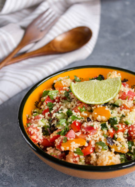 istock Tabbouleh salad: couscous and vegetables in the orange ceramic bowl on concrete surface. Fresh summer salad with lime slice, wooden spoon, fork and white linen towel 1243673011