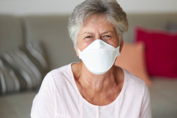 A senior citizen behind a face mask, cosmetic mask or surgical mask smiles at the camera A senior citizen behind a face mask, cosmetic mask or surgical mask smiles at the camera while sitting on the couch in the living room people laughing hard stock pictures, royalty-free photos & images