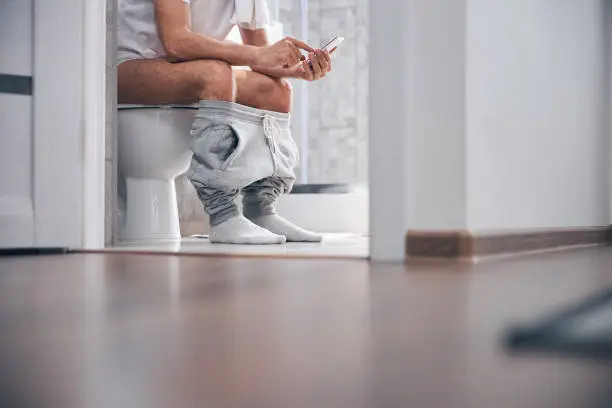Cropped photo of a Caucasian male with a cellular phone sitting on the toilet seat