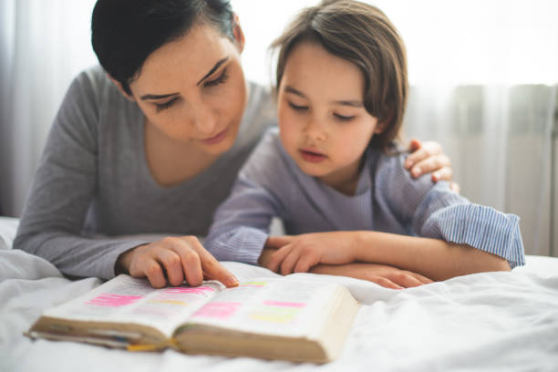 Mother and her daughter reading from bible and praying in their knees near the bed stock photo