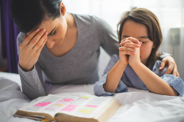Mother and her daughter reading from bible and praying in their knees near the bed stock photo