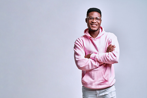 Handsome african american young man in glasses and pink hoodie standing with arms crossed on grey background. Copy space