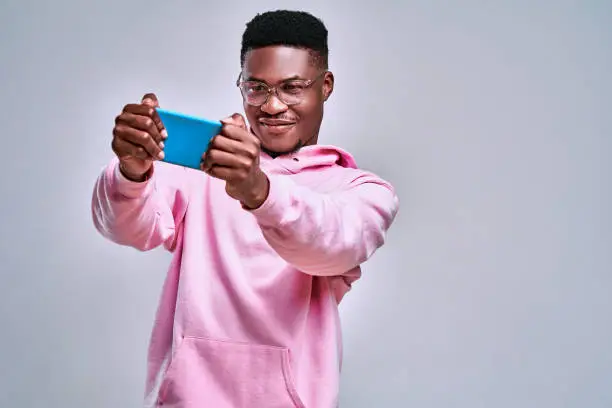 Photo of Portrait of a cheerful young african man dressed in pink hoodie playing games on mobile phone isolated over grey background
