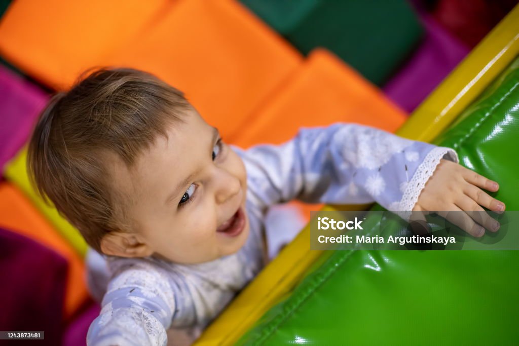 menina bonitinha em um vestido na piscina com cubos olha com sorriso e puxa a mão para cima para subir no playground no centro de brincadeiras infantis. vista superior - Foto de stock de Parque natural royalty-free menina bonitinha em um vestido na piscina com cubos olha com sorriso e puxa a mão para cima para subir no playground no centro de brincadeiras infantis. vista superior - Foto de stock de Parque natural royalty-free