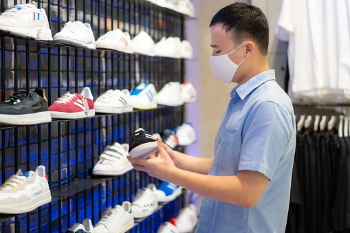 Asian man wearing mask over her face while choosing sport shoe for run at shopping mall for healthcare and prevention from coronavirus, Covid19 influenza in crowded place.