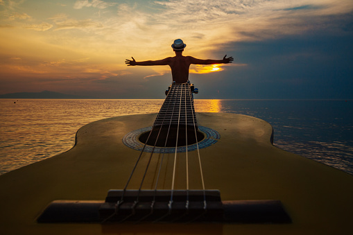 Young man with hat standing in sea in front of acoustic guitar at beautiful sunset, admiring nature and looking for an inspiration