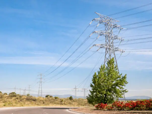 Huge electric tower next to a road and a roundabout decorated with red flowers and a blue sky with clouds in the background
