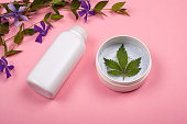 white bottles with body cosmetics with marijuana leaf on a pink background with violet wildflowers. skin care, beauty