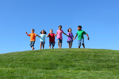 Group of multi-ethnic kids running together holding hands, on grass hill with blue sky