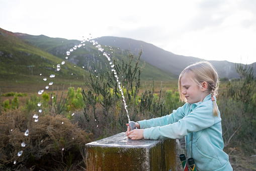A young girl tried to drink water from a water fountain while she is walking in the mountains. Water spraying from a tap