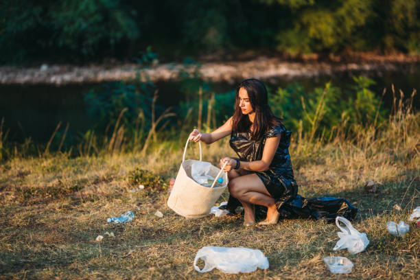 Young attractive woman in elegant dress made of garbage bags picking up garbage in nature.