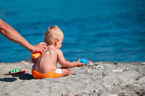 The male hand applies sunscreen to the baby is back. A child sits on the beach and plays with sand.
