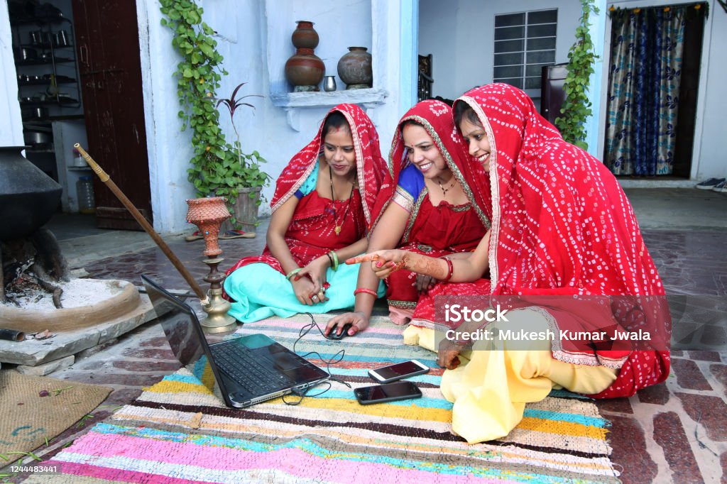 Traditional Indian young married women working in traditional kitchen on laptop Using technology in rural households. Women cook food and learn technology. Rural women using Laptop. India Stock Photo