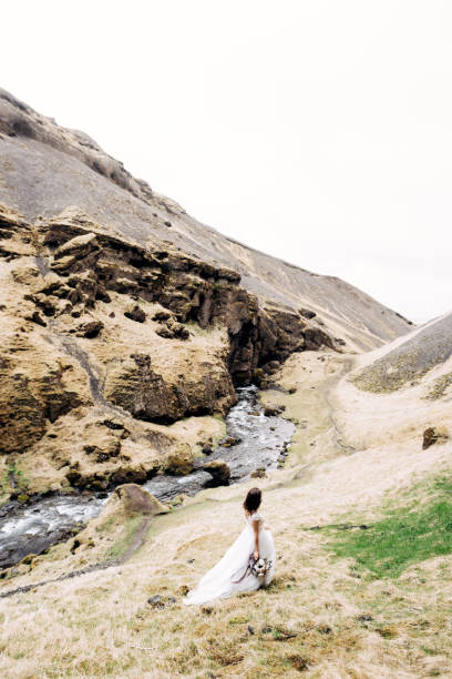 une mariée dans une robe blanche et un bouquet de fleurs dans ses mains marche le long des rives d’une rivière de montagne, près de la cascade de kvernufoss dans le sud de l’islande. - elope photos et images de collection