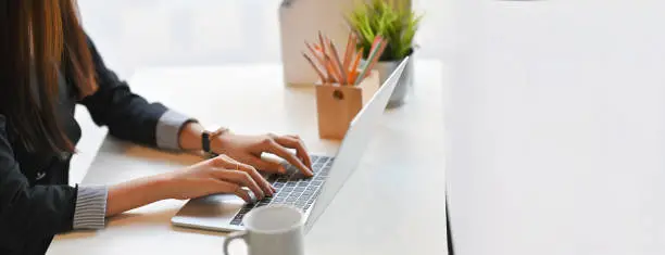 Photo of Cropped image of creative woman hands typing on a computer laptop that putting on a white working desk surrounded by a coffee cup, pencil holder, potted plant, and file folder.