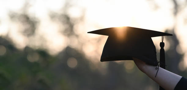 istock Cropped image of university student hand holding a graduation hat in hand over outdoors with sunset as background. 1244502773