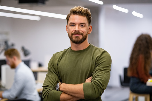 Portrait of proud businessman standing in creative modern office and looking at camera. Successful mid man entrepreneur standing with staff working in background. Satisfied young business man with crossed arms in advertising agency.