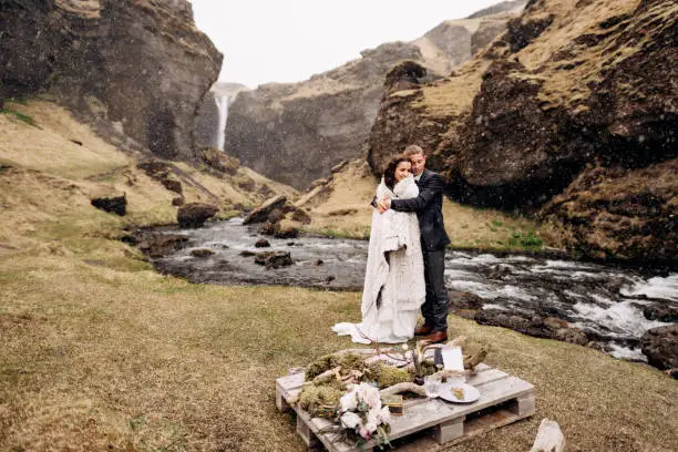 Photo of Destination Iceland wedding, near Kvernufoss waterfall. A wedding couple stands under a plaid near a mountain river. The groom hugs bride. They built an impromptu wedding table with decor and guitar