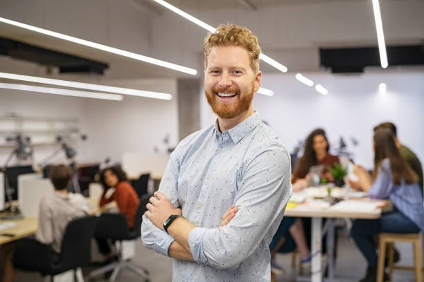 Successful business man smiling in office Happy successful businessman standing in office and looking at camera. Portrait of mid adult man entrepreneur standing in open space office with staff working in background. Confident business man with co-workers sitting at desk in background. creative space photos stock pictures, royalty-free photos & images