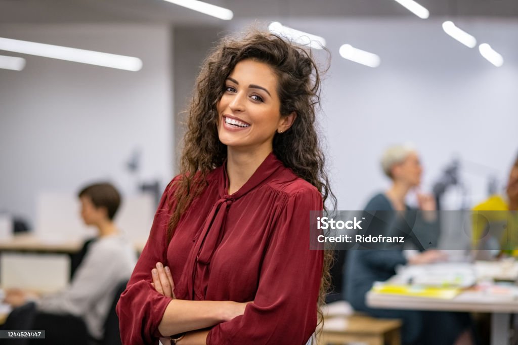Successful casual business woman smiling Successful businesswoman standing in creative office and looking at camera. Young latin woman entrepreneur in a coworking space smiling. Portrait of beautiful business woman standing in front of business team at modern agency with copy space. Women Stock Photo