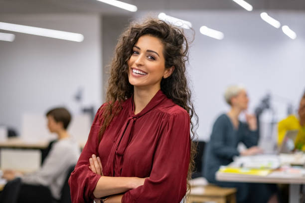 Successful casual business woman smiling Successful businesswoman standing in creative office and looking at camera. Young latin woman entrepreneur in a coworking space smiling. Portrait of beautiful business woman standing in front of business team at modern agency with copy space. businesswear stock pictures, royalty-free photos & images