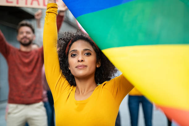 Proud woman in protest rally for lgbt rights Mixed race young woman at women empowerment strike holding rainbow flag. Proud multiethnic lesbian girl in rally to protest on equality for gay. Portrait of confident girl with group of activists outdoor during gay pride parade. bi black stock pictures, royalty-free photos & images