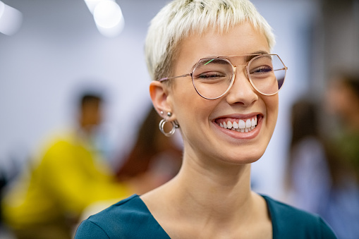 Successful business woman in modern office enjoy the positive result of her work. Close up face of creative stylish woman smiling and looking at camera. Portrait of trendy university student laughing with classmates working in background.