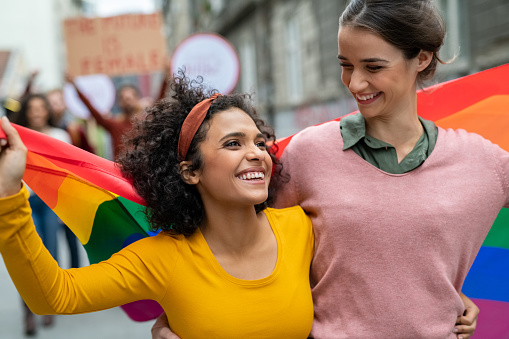 Young cheerful women on street enjoying holding gay pride flag during protest. Smiling multiethnic women enjoying victory after march on street for lgbt rights. Diversity, tolerance and gender identity concept.