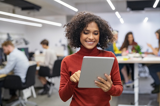 Happy multiethnic businesswoman with curly hair using digital tablet in office. Successful young woman working on laptop. Smiling casual mixed race business woman standing in workplace with colleagues sitting at desk in background.