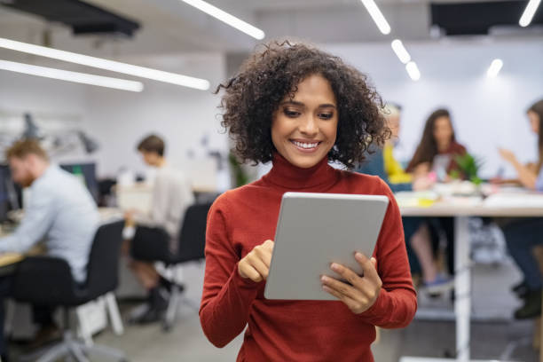 Multiethnic young businesswoman using digital tablet Happy multiethnic businesswoman with curly hair using digital tablet in office. Successful young woman working on laptop. Smiling casual mixed race business woman standing in workplace with colleagues sitting at desk in background. creative space photos stock pictures, royalty-free photos & images