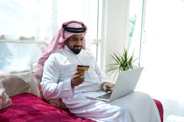 Saudi Arab man using laptop Man in Saudi Arabia is using laptop and doing online purchase and banking white sitting and relaxing at home and maintaining social distance al madinah photos stock pictures, royalty-free photos & images