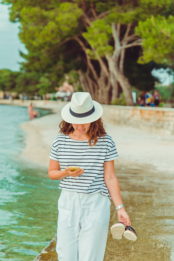 woman in white clothes and fedora hat walking by sea beach summer time