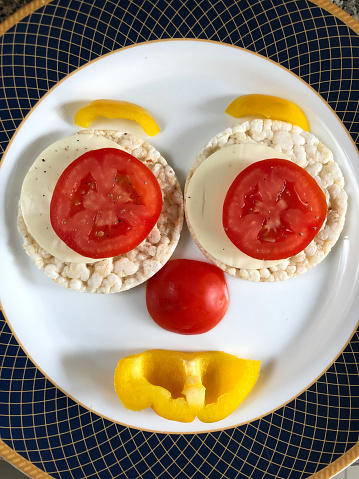 Stock photo of healthy rice cakes topped up with cheese, tomato and yellow bell pepper in ceramic plate, healthy snack food plated as smiley face for children to look appetising. rice cake made with brown puffed rice good for health and help in weight loss