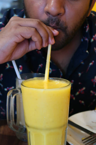 Stock photo showing unrecognised man drinking mango milkshake, smoothie with plastic straw, man with beard sucking / sipping  milkshake, healthy eating five a day fruit and vegetable portions, glass of mango juice seasonal drinks made with milk, ripe mango and nuts