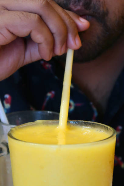 Stock photo showing unrecognised man drinking mango milkshake, smoothie with plastic straw, man with beard sucking / sipping  milkshake, healthy eating five a day fruit and vegetable portions, glass of mango juice seasonal drinks made with milk, ripe mango and nuts