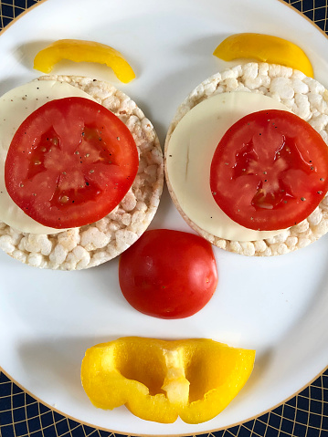 Stock photo of healthy rice cakes topped up with cheese, tomato and yellow bell pepper in ceramic plate, healthy snack food plated as smiley face for children to look appetising. rice cake made with brown puffed rice good for health and help in weight loss