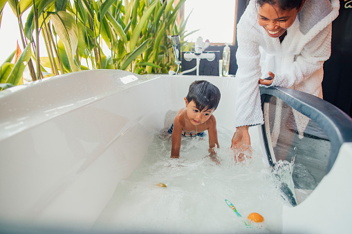An Indonesian boy playing in the bath at home in Bali, Indonesia. His mother is standing outside the bath while splashing the water with her hand.