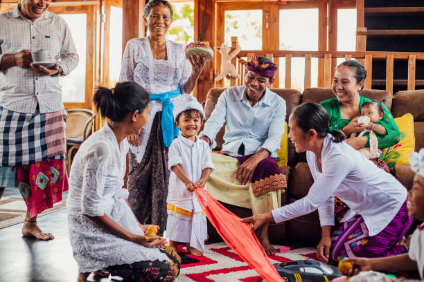 A wide shot of an Indonesian family and their babysitter together in the living room. The youngest son is playing a game with material while the nanny helps. They are at home in Bali, Indonesia.
