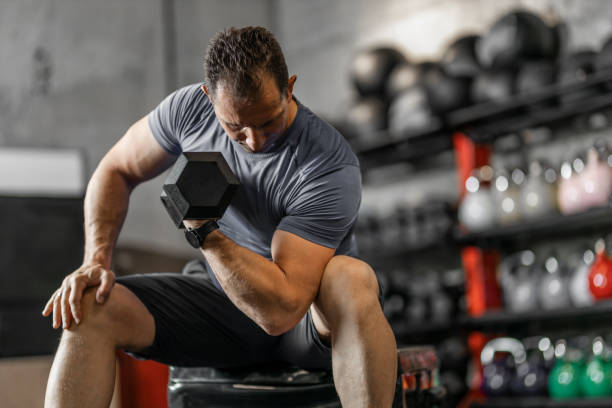 istock Muscular guy in sportswear lifting dumbbell while sitting on bench at cross training gym. Mature athlete using dumbbell during a workout. Strong man under physical exertion pumping up bicep muscle with heavy weight. 1244729137