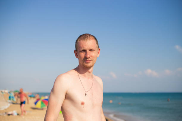 a young man with a bare torso is standing on the seashore and squinting his eyes in displeasure