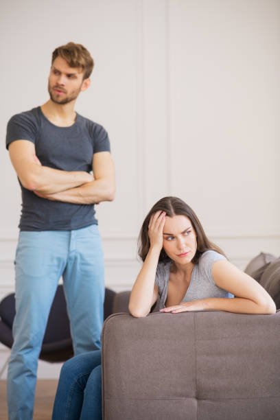 istock Young bearded man standing with arms clasped while his wife sitting with depressed look 1244754572