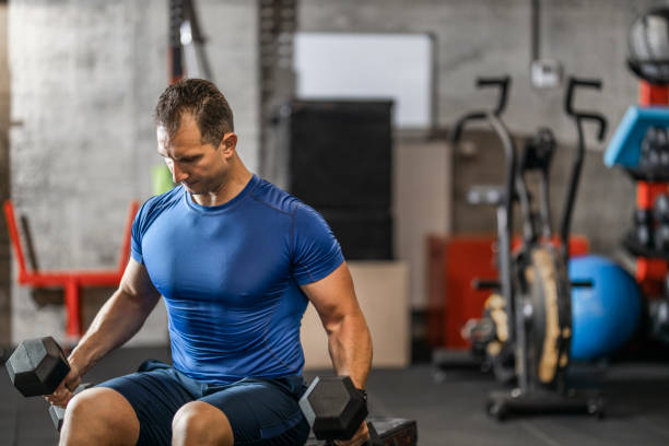 istock Muscular guy in sportswear lifting dumbbell while sitting on bench at cross training gym. Mature athlete using dumbbell during a workout. Strong man under physical exertion pumping up bicep muscle with heavy weight. 1244760120