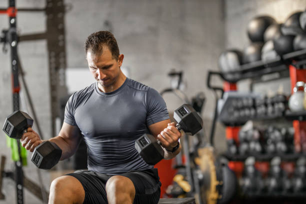 istock Muscular guy in sportswear lifting dumbbell while sitting on bench at cross training gym. Mature athlete using dumbbell during a workout. Strong man under physical exertion pumping up bicep muscle with heavy weight. 1244761138